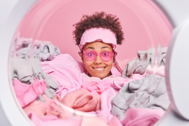 Foto grátis mulher de cabelo encaracolado morde os lábios e olha alegremente para a câmera feliz por terminar o trabalho doméstico usa óculos de sol da moda varando a cabeça através de uma pilha de poses de roupa suja na máquina de lavar