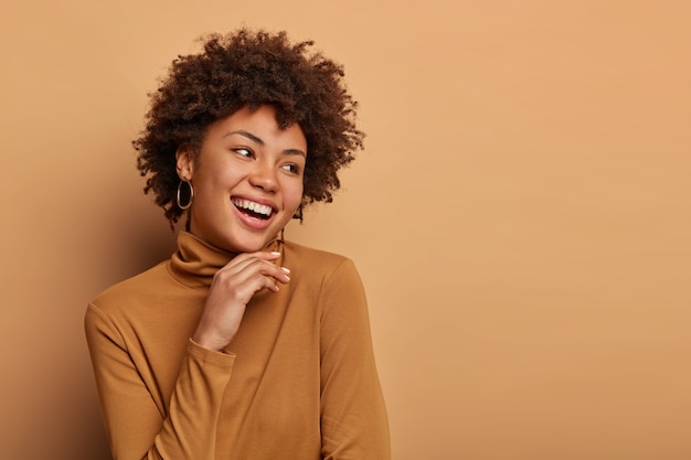 Mulher de cabelo encaracolado olha alegremente para o lado, toca o queixo suavemente, tem um largo sorriso dentu