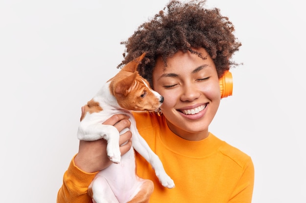 Mulher de cabelo encaracolado positiva sente-se cuidada e responsável segurando um pequeno cão de raça junto em casa ouve música em fones de ouvido sem fio fecha os olhos com ternura isolada sobre a parede branca