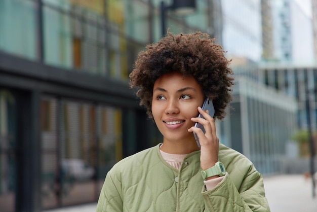 Foto grátis mulher de cabelo encaracolado sorridente tem conversa telefônica gosta de tarrifs móveis e conexão ligando para amigo usa jaqueta focada em algum lugar posa ao ar livre contra fundo desfocado comunicação