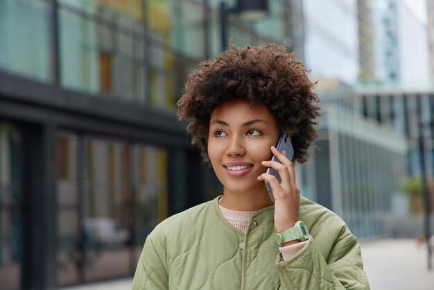 Mulher de cabelo encaracolado sorridente tem conversa telefônica gosta de tarrifs móveis e conexão ligando para amigo usa jaqueta focada em algum lugar posa ao ar livre contra fundo desfocado Comunicação