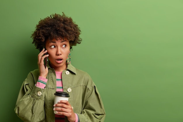 Foto grátis mulher de cabelo encaracolado surpresa e emocional segura o smartphone perto da orelha, parece com uma expressão chocada, segura uma xícara descartável de café, usa roupas da moda,