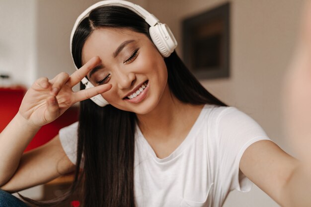 Mulher de cabelo escuro usando fones de ouvido mostra o símbolo da paz e sorri com os olhos fechados