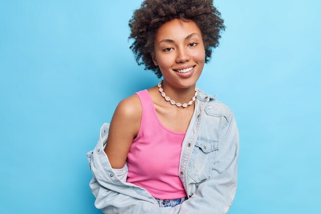 Mulher de cabelo muito encaracolado usa camiseta rosa e colar de jaqueta jeans mostra sorrisos de ombro nu alegremente isolados na parede azul