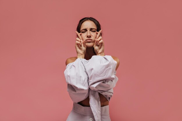 Mulher de cabelo preta na moda na blusa listrada leve e saia legal cruzando os dedos e posando com os olhos fechados no pano de fundo rosa