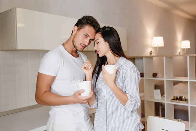 Mulher de cabelo preto com sono na cozinha com o namorado bebendo uma bebida quente