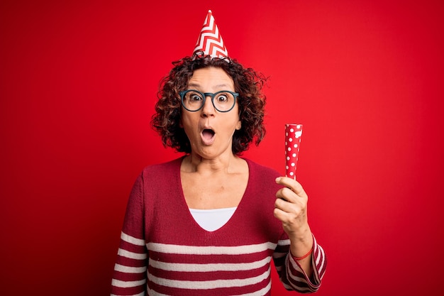 Foto grátis mulher de cabelos cacheados de meia-idade usando chapéu engraçado de aniversário segurando trompete de festa na celebração assustada em estado de choque com uma cara de surpresa com medo e animada com expressão de medo