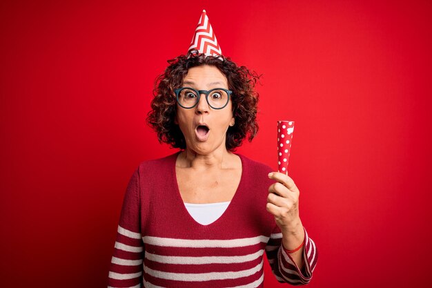 Mulher de cabelos cacheados de meia-idade usando chapéu engraçado de aniversário segurando trompete de festa na celebração assustada em estado de choque com uma cara de surpresa com medo e animada com expressão de medo