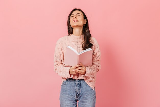 Mulher de cabelos escuros alegre ri enquanto lê o livro. Retrato de uma menina de cabelos escuros numa camisola clara no fundo rosa.