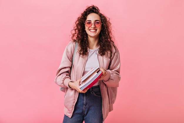 Mulher de cabelos escuros em jeans e jaqueta rosa sorrindo e segurando livros