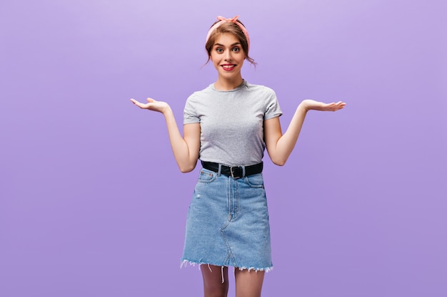 Mulher de cabelos escuros em roupas jeans encolhendo os ombros. menina bonita encantadora com lábios cor de rosa em t-shirt cinza posando para a câmera em fundo isolado.