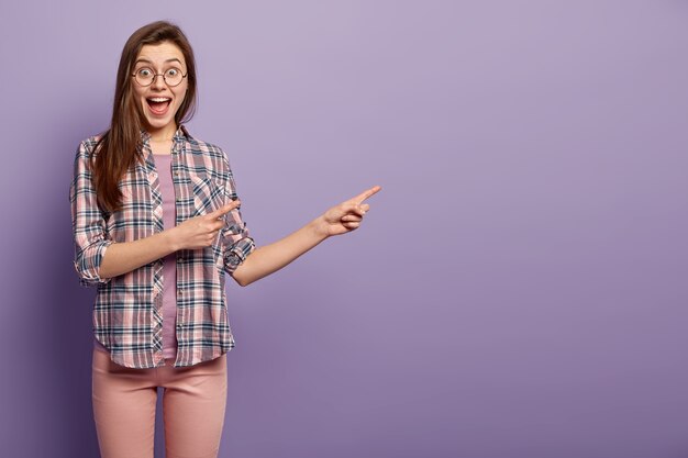 Mulher de cabelos escuros encantada em camisa xadrez, sorri amplamente, indica no espaço da cópia