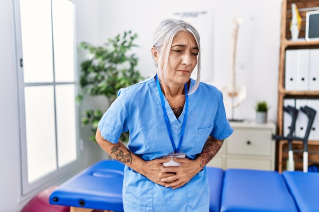 Mulher de cabelos grisalhos de meia-idade vestindo uniforme de fisioterapeuta na clínica médica com a mão no estômago porque náusea doença dolorosa sensação de mal-estar conceito de dor