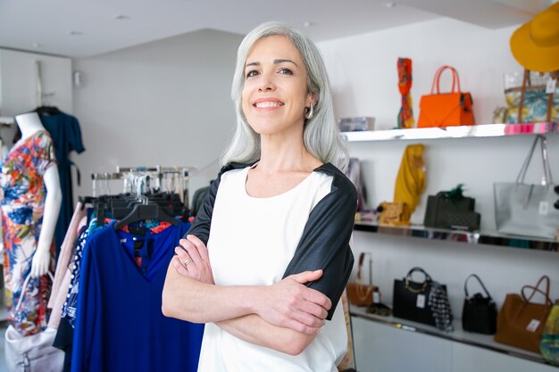 Mulher de cabelos louros caucasiana alegre em pé com os braços cruzados perto do rack com vestidos na loja de roupas, olhando para a câmera e sorrindo. Cliente de boutique ou conceito de assistente de loja