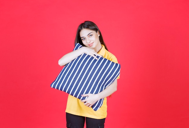 Mulher de camisa amarela segurando um travesseiro azul com listras brancas e parece pensativa.