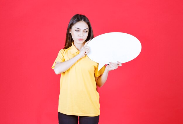 mulher de camisa amarela segurando uma placa de informações oval e parece confusa e pensativa.