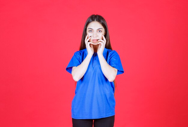 mulher de camisa azul em pé na parede vermelha e parece emocionada e apavorada.