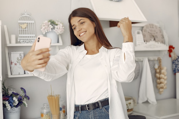 Mulher de camisa branca em pé na cozinha e fazendo uma selfie