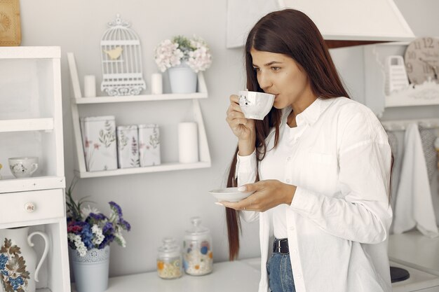 Mulher de camisa branca em pé na cozinha e tomando um café