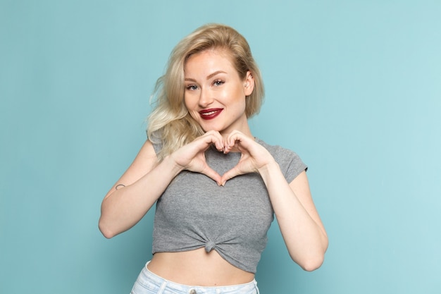 Mulher de camisa cinza e jeans azul brilhante posando com um sorriso e mostrando o sinal do coração