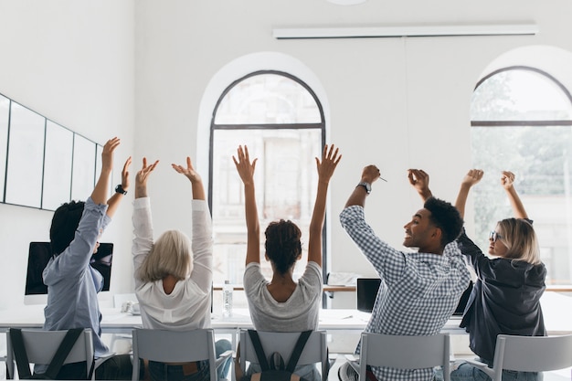 Mulher de camisa formal com cabelo loiro, acenando com as mãos, sentada entre colegas de trabalho na sala de conferências grande luz. Foto da parte de trás de gerentes cansados, alongando-se durante uma reunião no escritório.