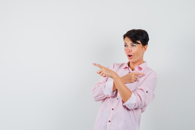 Mulher de camisa rosa, apontando para algo longe e olhando espantada, vista frontal.