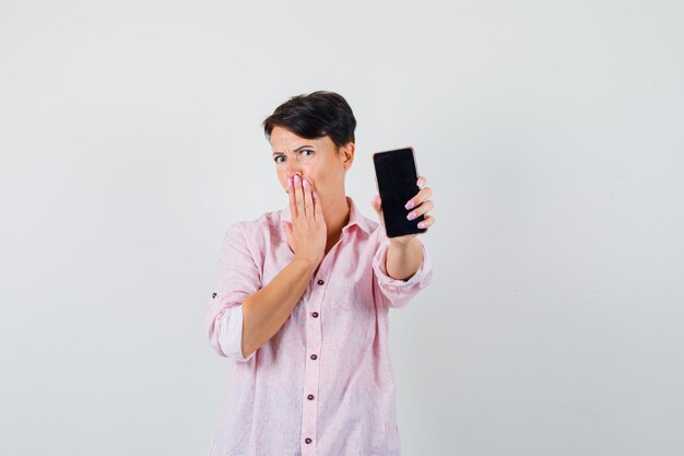 Mulher de camisa rosa, mostrando o celular e parecendo surpresa, vista frontal.