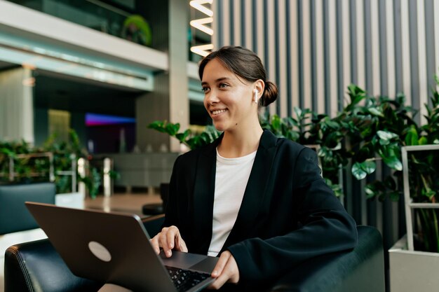 Mulher de casaco e camisa com cabelo recolhido com sorriso trabalhando no laptop e ouvindo música com fones de ouvido sem fio