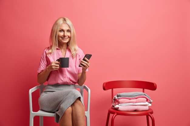 Foto grátis mulher de cinquenta anos satisfeita segura um telefone celular e uma caneca de chá, surf nas redes sociais, enquanto passa o tempo livre em casa, senta-se em uma cadeira confortável sozinha e gosta de comunicação online. conceito de estilo de vida