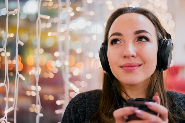 Foto grátis mulher de close-up usando fones de ouvido segurando xícara perto de luzes de natal