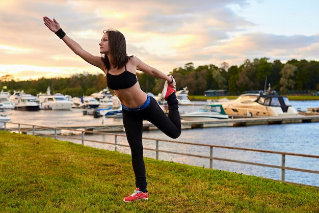Mulher de esporte fitness fazendo alongamento com as pernas durante o treino de cross training ao ar livre. Mulher em uma blusa preta e meia-calça sob o sol forte.