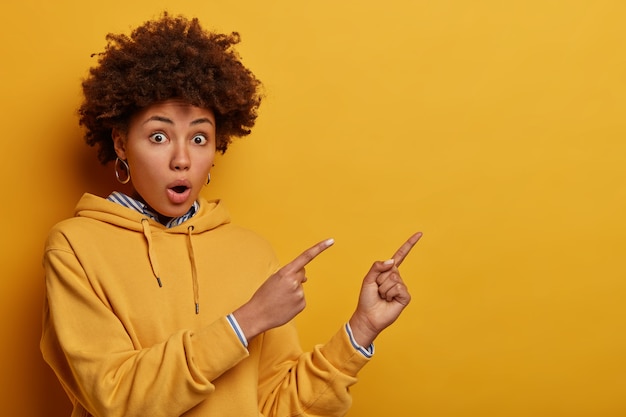 Foto grátis mulher de etnia adulta surpresa e intrigada com penteado afro apontando no canto superior direito, mostra cartaz ou banner publicitário com grande admiração, chocada com o preço alto, vestida com moletom amarelo