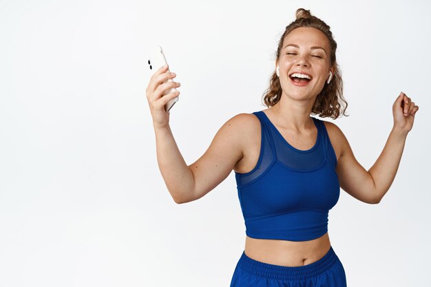 Mulher de fitness feliz dançando em fones de ouvido sem fio e roupas esportivas. Atleta de garota segurando smartphone e rindo, de pé sobre fundo branco