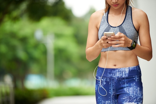 Mulher de forma irreconhecível em esportes top e leggings, com fones de ouvido, usando smartphone