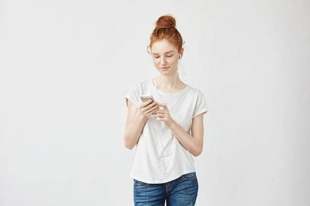 Mulher de gengibre com sardas em fones de ouvido, sorrindo, olhando para o telefone.