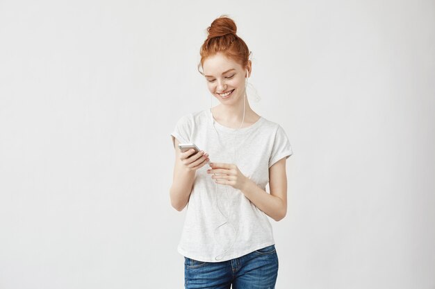 Mulher de gengibre com sardas em fones de ouvido, sorrindo, olhando para o telefone.