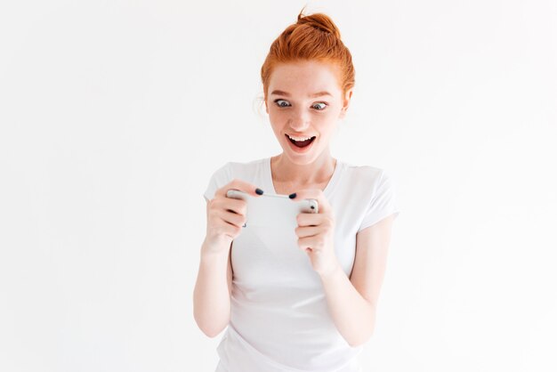 Mulher de gengibre feliz surpresa em t-shirt jogando em seu smartphone