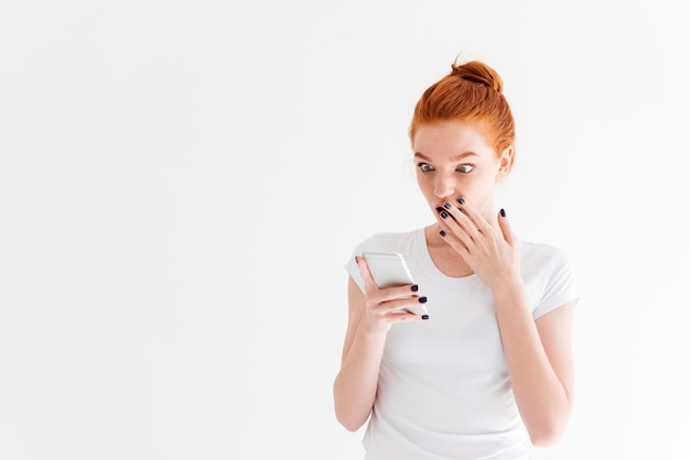 Mulher de gengibre surpresa em t-shirt usando seu smartphone