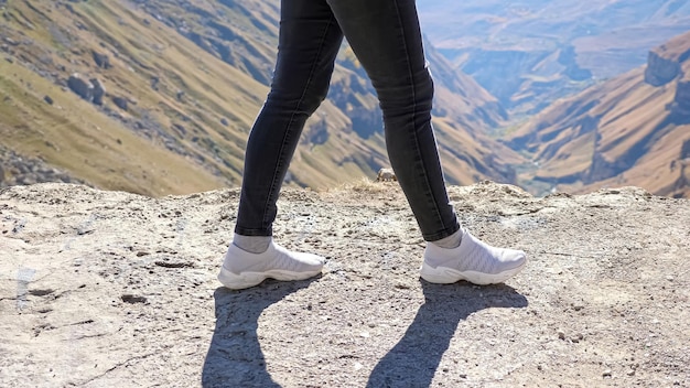 Mulher de jeans preto e tênis cinza claro caminha ao longo da borda de um penhasco rochoso contra montanhas distantes e desfiladeiro profundo.