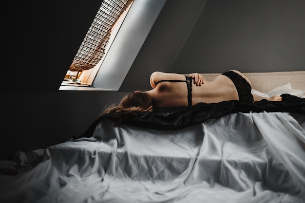 Mulher de lingerie preta encontra-se na cama cinza antes da janela