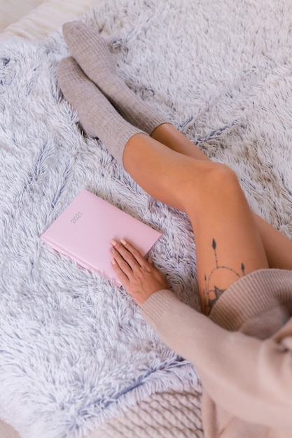 Mulher de meia de lã e blusa com caderno rosa assinam 2021, grande tatuagem no quadril. Mulher sente-se na cama em casa, no quarto.