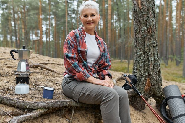 Mulher de meia idade alegre ativa sentada sob uma árvore com equipamento de acampamento fervendo água para chá no fogão a gás, tendo uma pequena pausa durante a caminhada de longa distância. Pessoas, aventura, viagens e caminhadas
