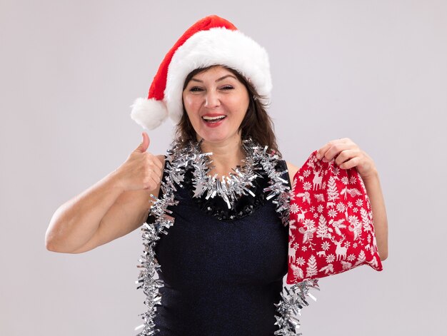 Mulher de meia-idade alegre com chapéu de Papai Noel e guirlanda de ouropel no pescoço segurando um saco de presente de Natal, olhando para a câmera mostrando o polegar isolado no fundo branco