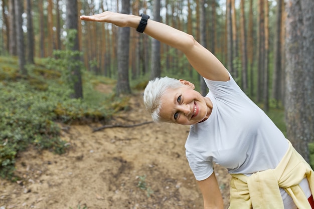 Mulher de meia-idade ativa e energética aquecendo o corpo antes de correr, posando contra pinheiros