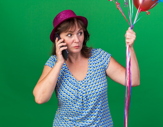 Mulher de meia-idade com chapéu de festa segurando balões coloridos, parecendo confusa enquanto fala no celular, comemorando a festa de aniversário em pé sobre a parede verde