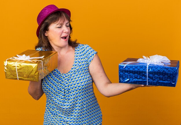 Mulher de meia-idade com chapéu de festa segurando presentes de aniversário, olhando para eles, feliz e alegre, sorrindo amplamente