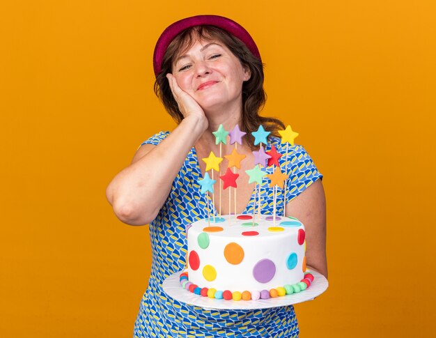 Mulher de meia-idade com chapéu de festa segurando um bolo de aniversário feliz e positiva sorrindo alegremente