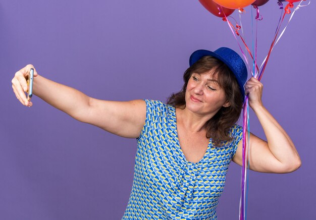 Mulher de meia-idade com chapéu de festa segurando um monte de balões coloridos, feliz e alegre fazendo selfie usando smartphone para celebrar a festa de aniversário em pé sobre a parede roxa