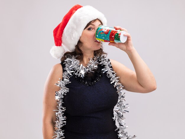 Mulher de meia-idade com chapéu de papai noel e guirlanda de ouropel no pescoço, olhando para a câmera, bebendo café em um copo de plástico de natal isolado no fundo branco