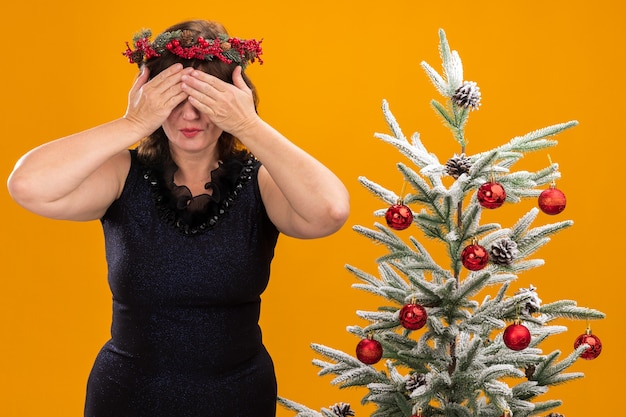 Mulher de meia-idade com coroa de natal na cabeça e guirlanda de ouropel no pescoço em pé perto da árvore de natal decorada